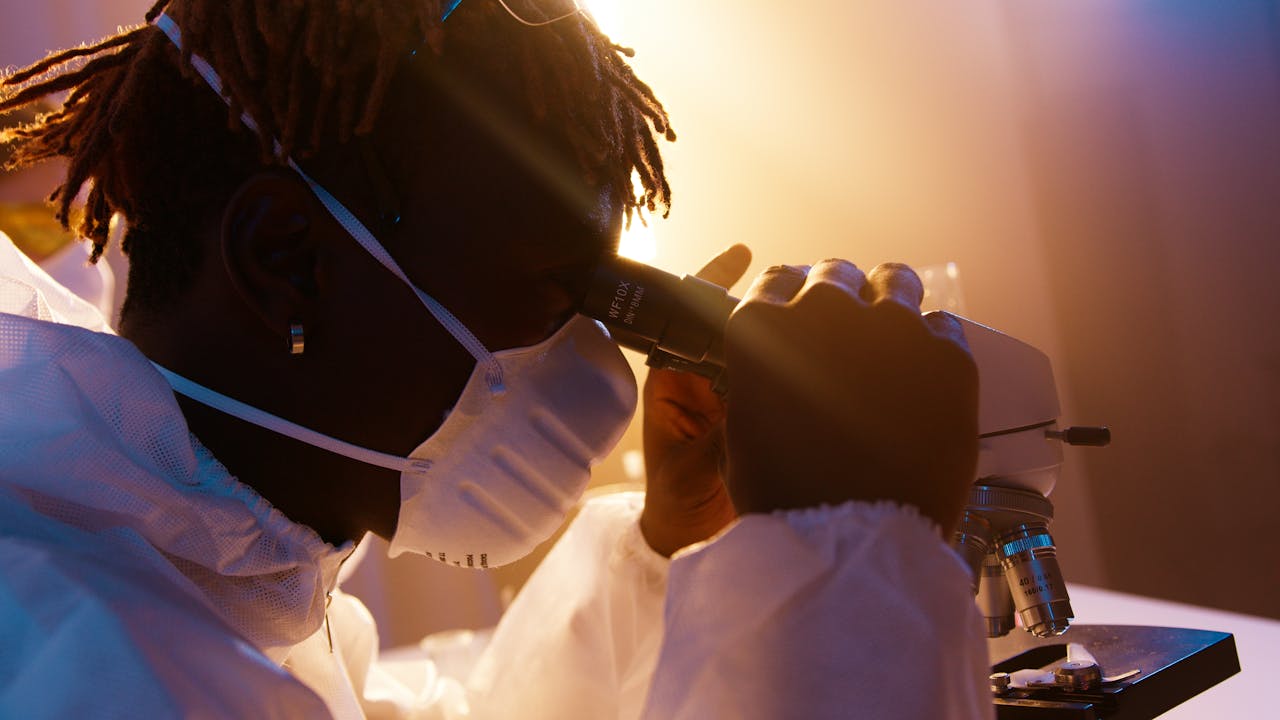 Scientist wearing protective gear examining samples under a microscope in a laboratory.