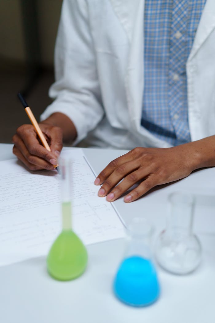 A student in a lab coat writes notes beside flasks filled with colored liquids in a laboratory setting.
