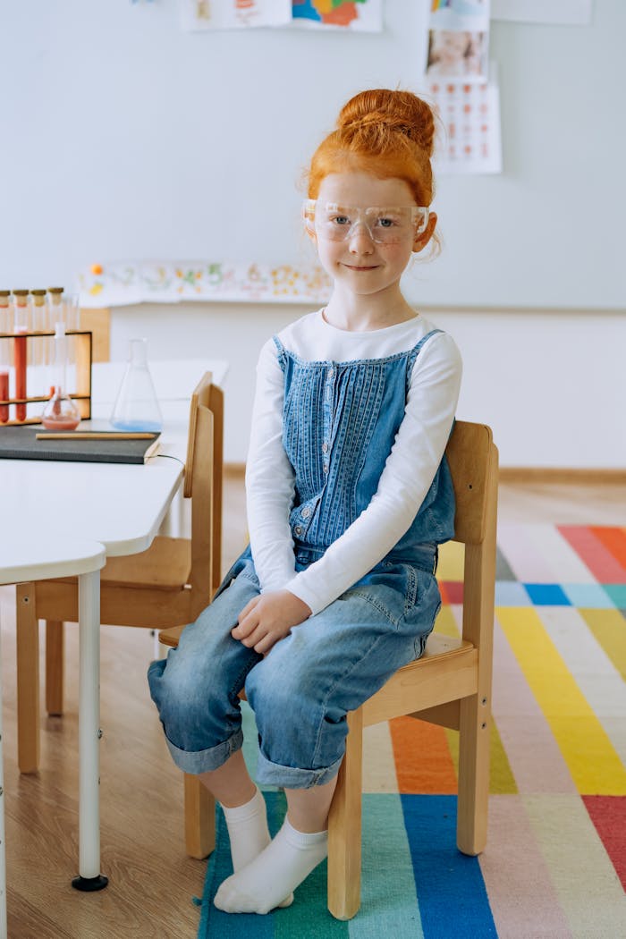 Child in classroom setting wearing safety glasses, sitting on wooden chair.